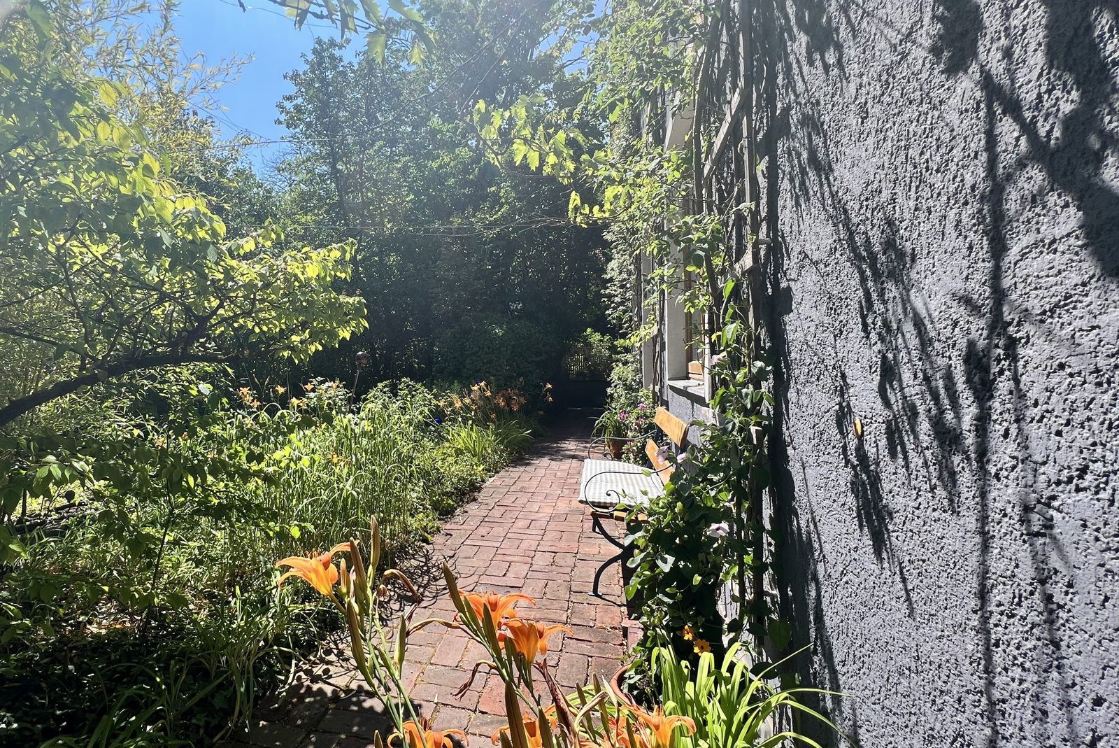 Sonniger Innenhofgarten mit Ziegelweg, Bank und blühenden Lilien an der Hauswand